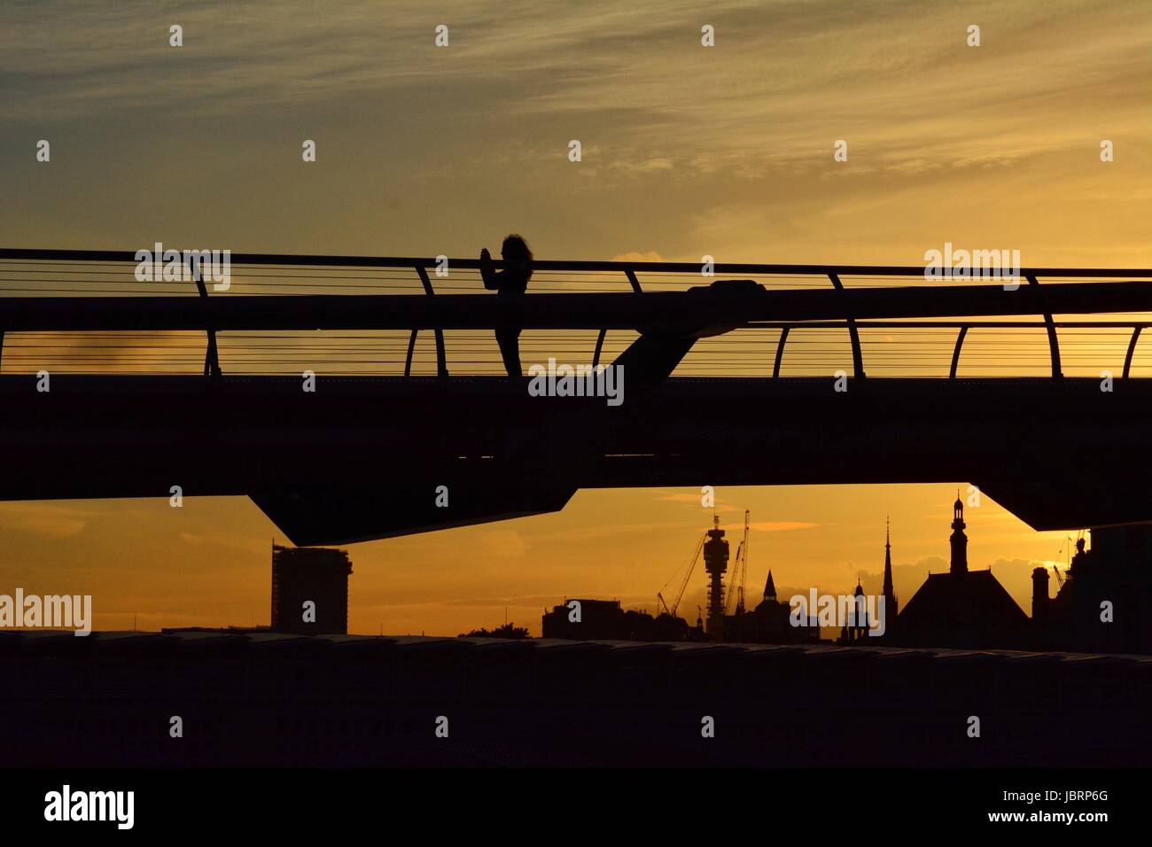 London, UK. 12th June 2017. A woman takes a photograph and is silhouetted against the sky as the sun sets behind the Millennium Bridge in London, UK. The BT tower is visible in the skyline beyond. Credit: Patricia Phillips/Alamy live news Stock Photo