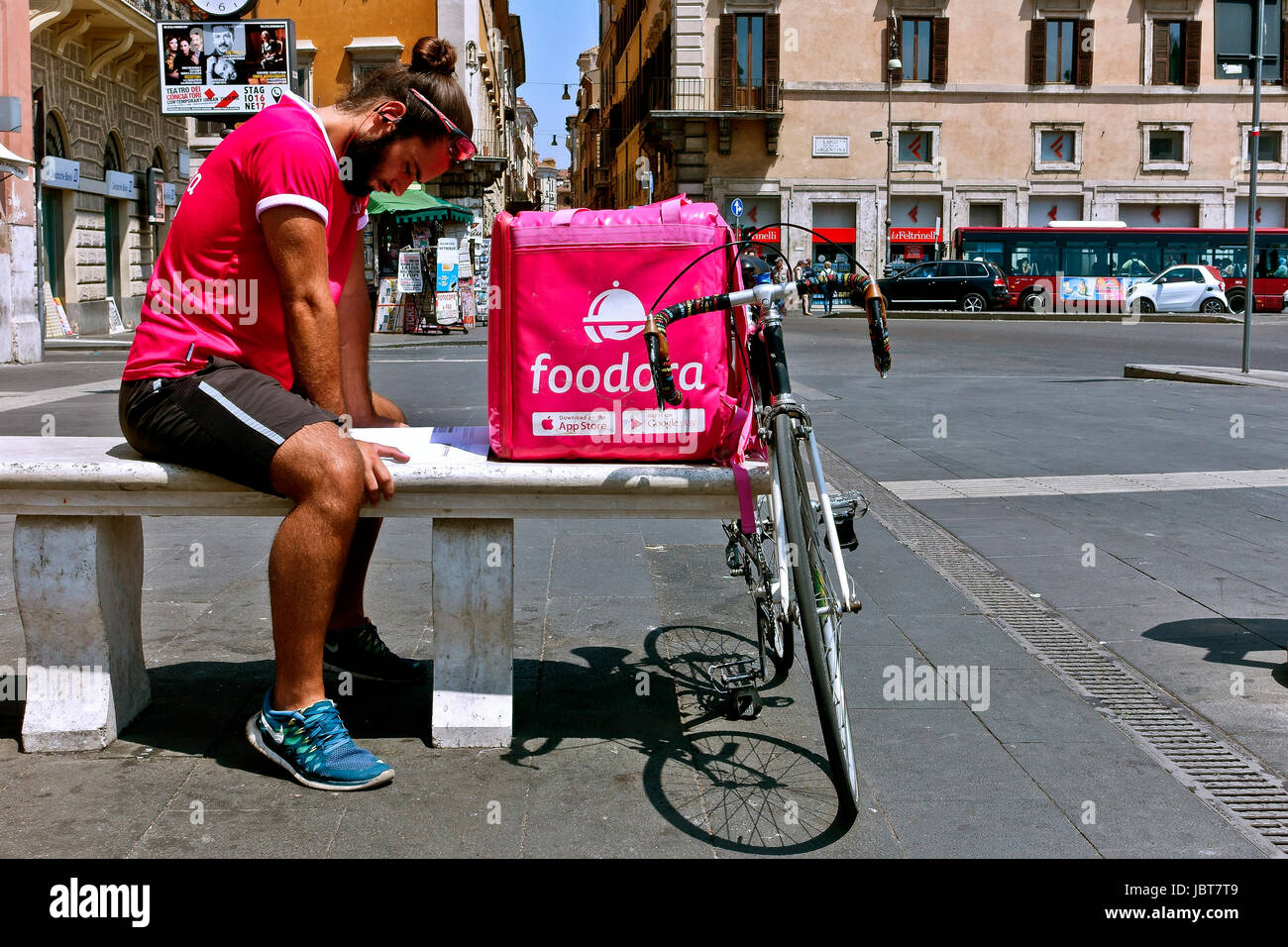 Delivery boy wearing a pink tshirt, resting on a bench and studying while waiting for a delivery request call. Pink delivery courier box. Rome, Italy Stock Photo