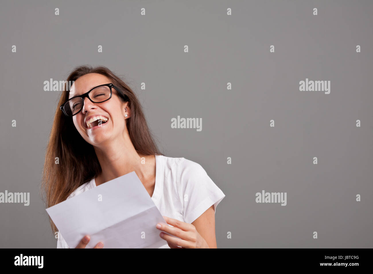 brunette girl with glasses laughing about news she received on a white document on a neutral gray faded background Stock Photo