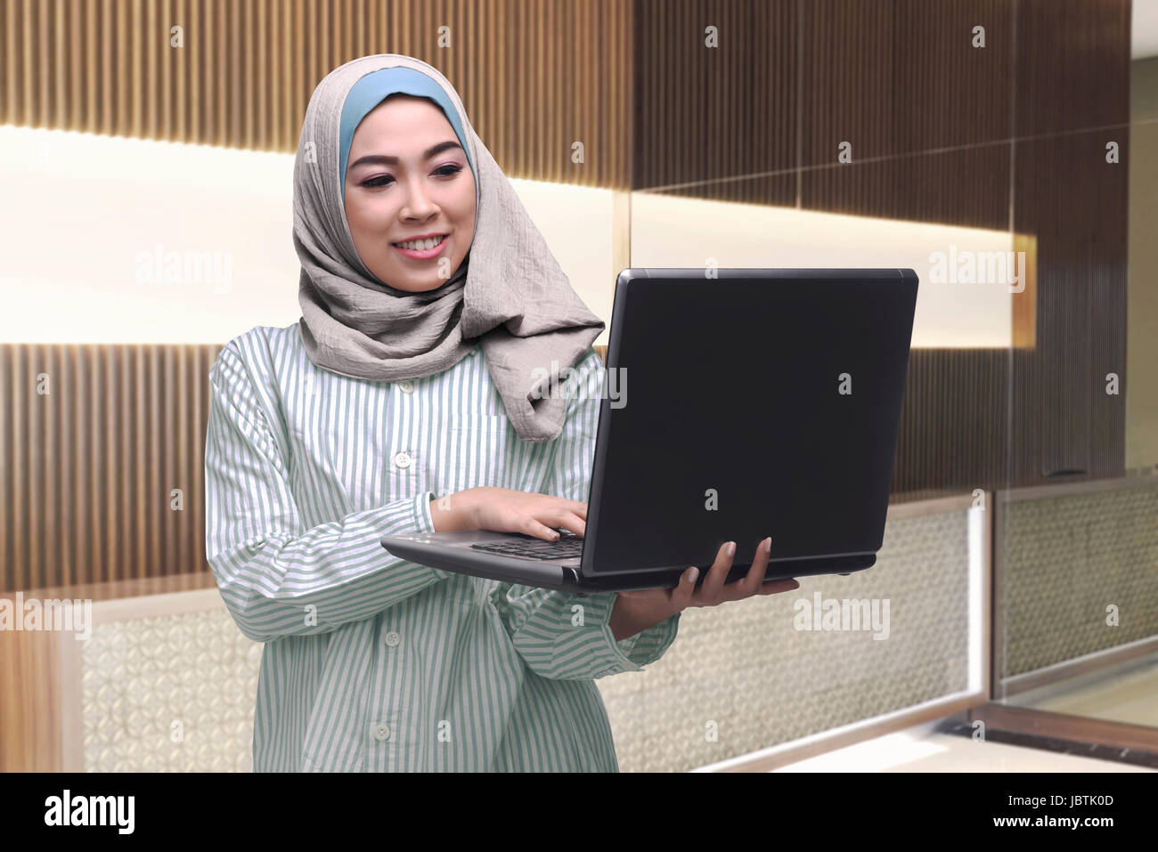 Beautiful asian muslim woman standing while typing on laptop inside her ...