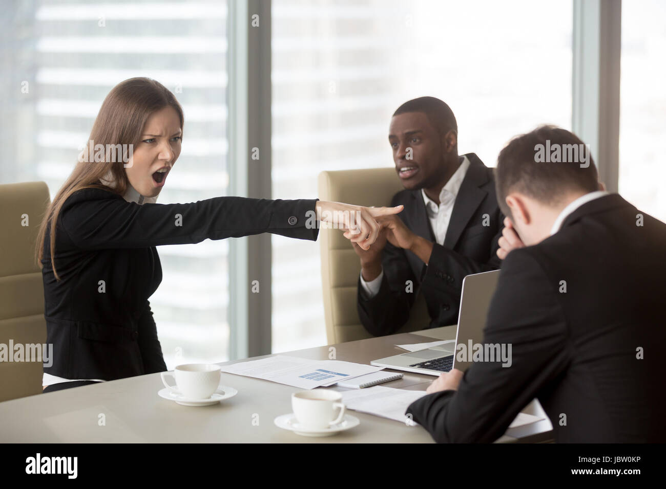 Angry female furious boss scolding employee at mixed-race team meeting, firing dismissing depressed office worker for failure, bad work results, being Stock Photo