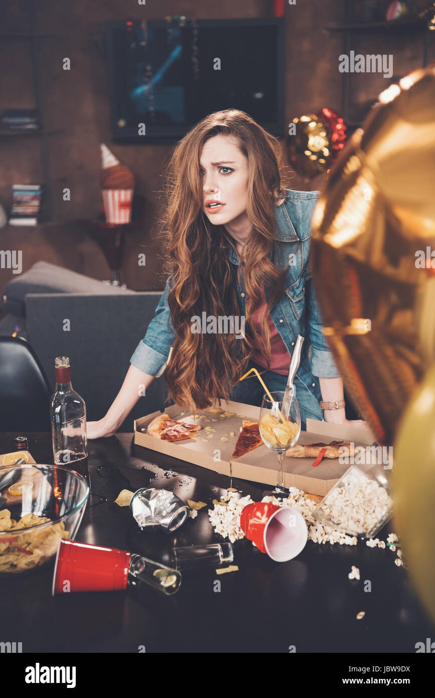 Depressed young woman with hangover leaning at messy table after party ...