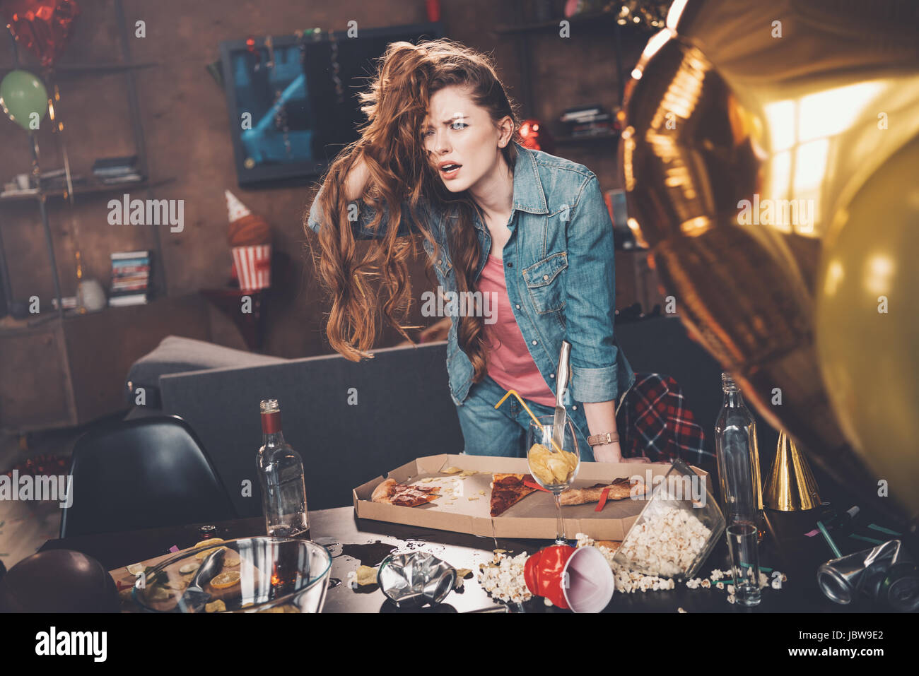 Depressed young woman with hangover leaning at messy table after party ...