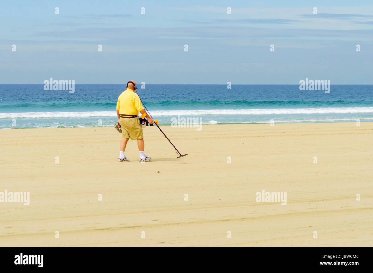 An old man walking along the coast holding a portable metal detector on