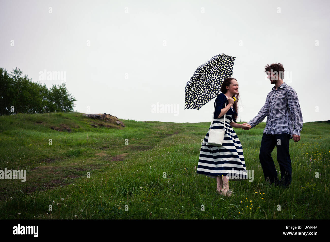 Couple standing in field, holding hands, young woman holding umbrella Stock Photo
