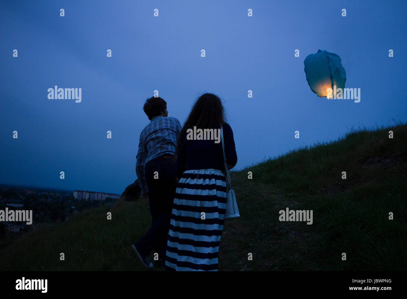 Couple walking up grassy hill at dusk, watching sky lantern in air, rear view Stock Photo