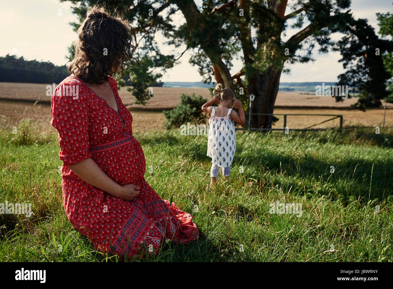 Pregnant woman sitting in field watching toddler daughter Stock Photo
