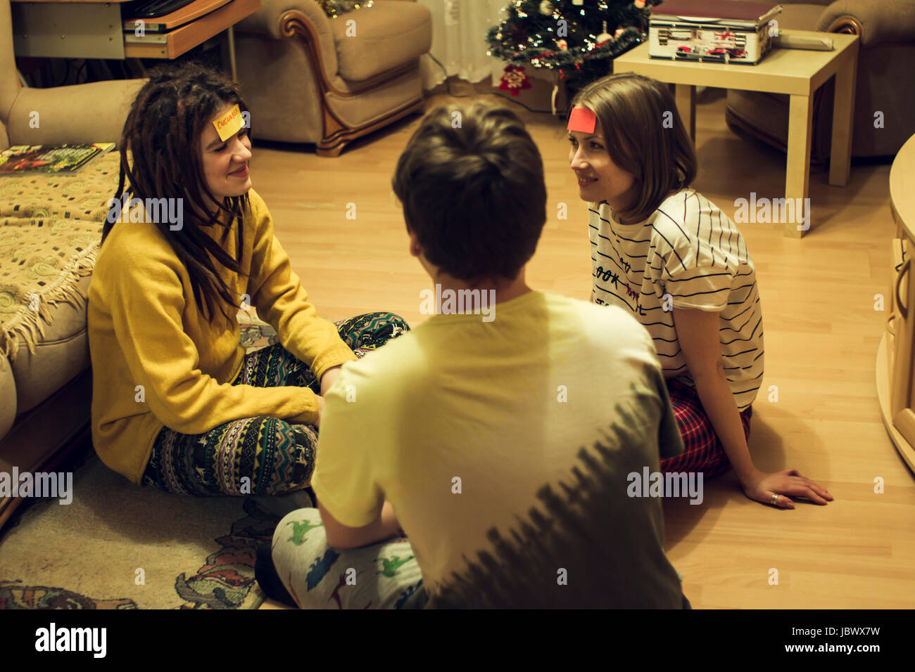 Three friends, sitting at home, playing game, stick notes stuck to their heads Stock Photo