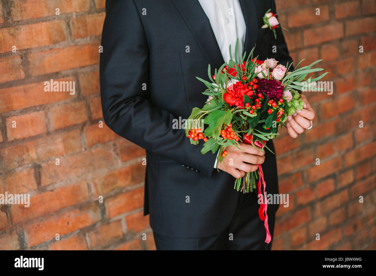 Elegant Man Dressed In A Modern Black Suit And White Shirt Holding In Hand A Wedding Flowers Bouquet From Red Carnation Close Up Color Vertical Image Of Male Hands Holding Bunch Of