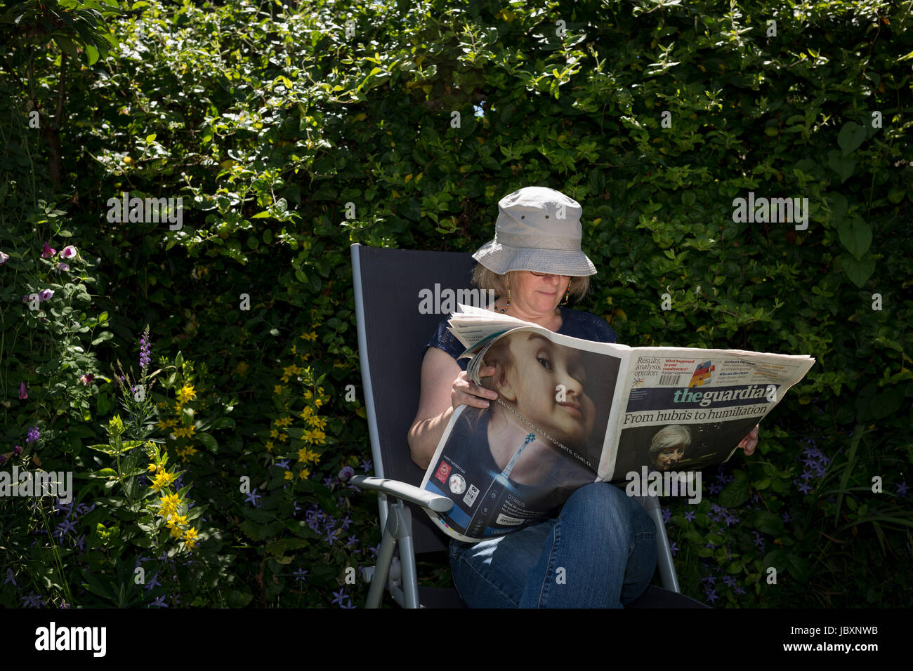 A middle-aged lady reads the Guardian newspaper featuring a toothbrush ad and Prime Minister Theresa May with the headline 'Hubris and Humiliation' in a shady spot of her summer garden, Two days after the 2017 general election, on 10th June 2017, in London, England. Stock Photo