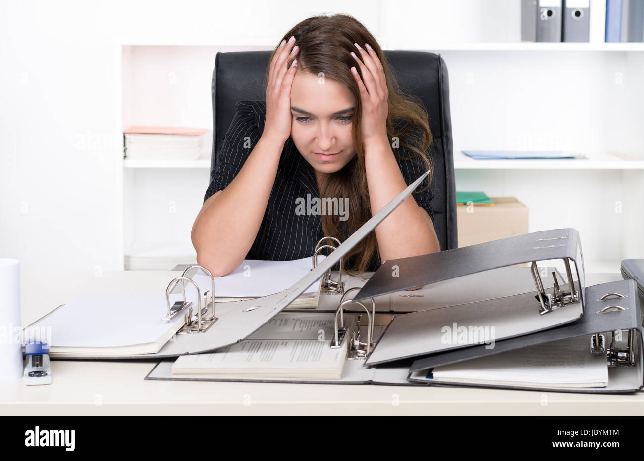 Eine junge, frustrierte Frau sitzt vor einem Haufen von Akten am Schreibtisch im Büro. Im Hintergrund steht ein Regal. Die Frau schaut nach unten. Stock Photo