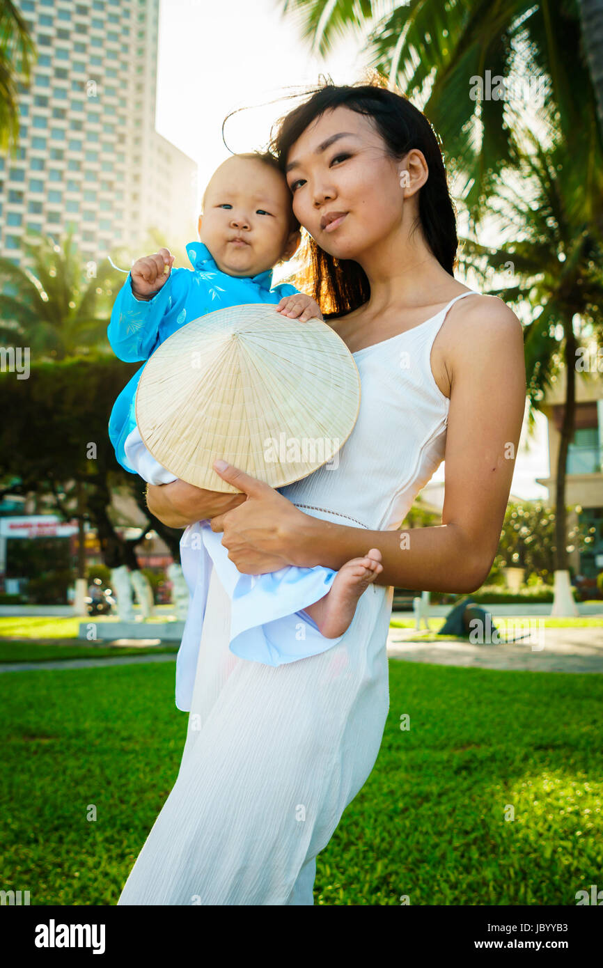 A mother holds her little daughter in traditional vietnam clothers aodai in her arms on the beach near palms Stock Photo