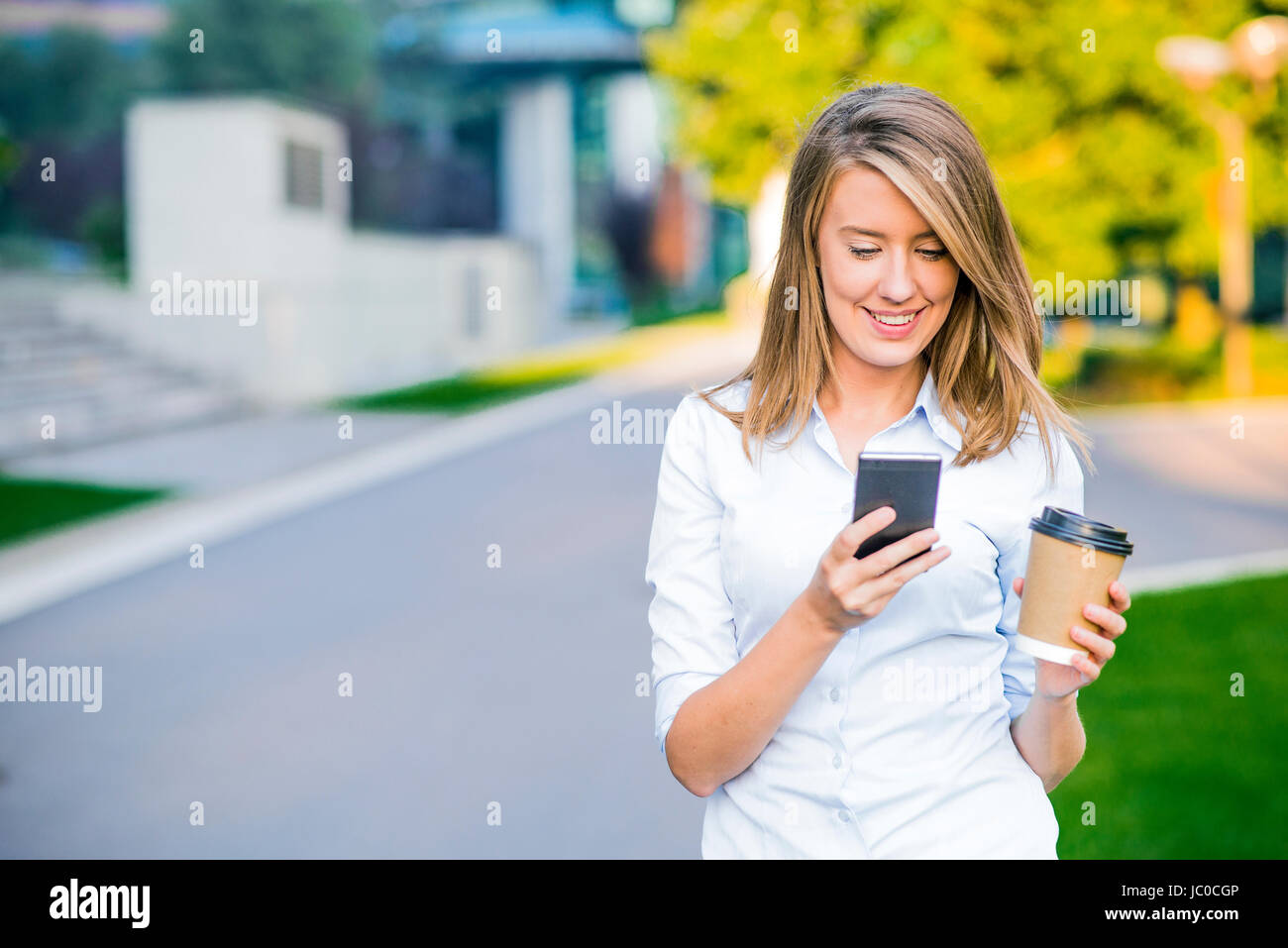 Young smart professional woman reading using phone. Female businesswoman reading news or texting sms on smartphone while drinking coffee on break from Stock Photo