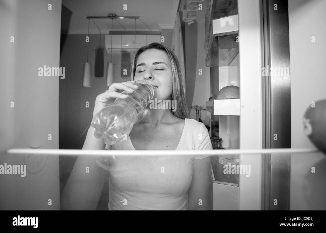 Black and white portrait of beautiful smiling woman taking water from fridge and drinking it. View from inside of open refrigerator Stock Photo