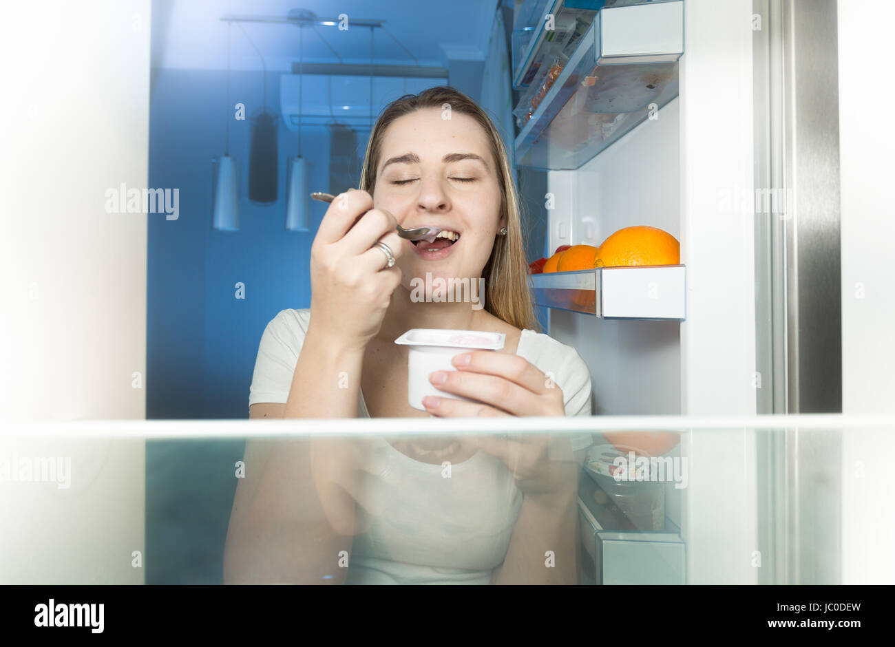 View from inside of refrigerator on young woman eating yogurt at night