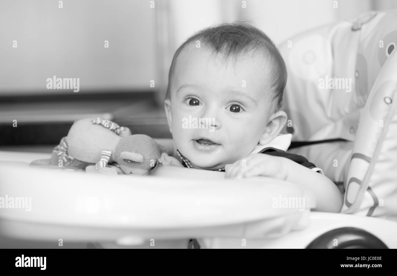 Black and white portrait of cute smiling baby boy sitting in highchair at kitchen Stock Photo