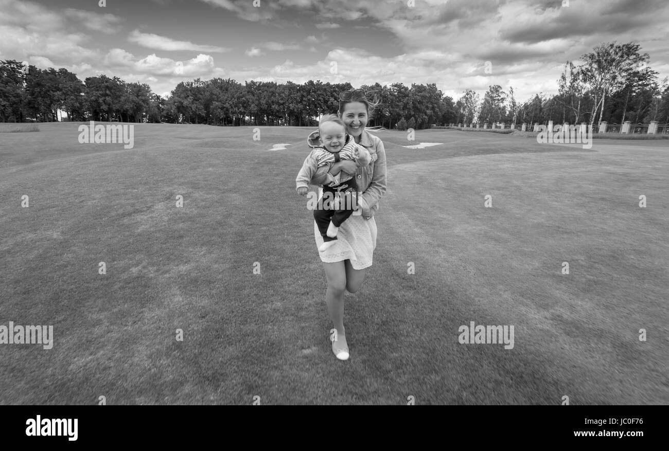 Black and white image of happy young mother running on grass at park with her 9 months old baby boy on hands Stock Photo