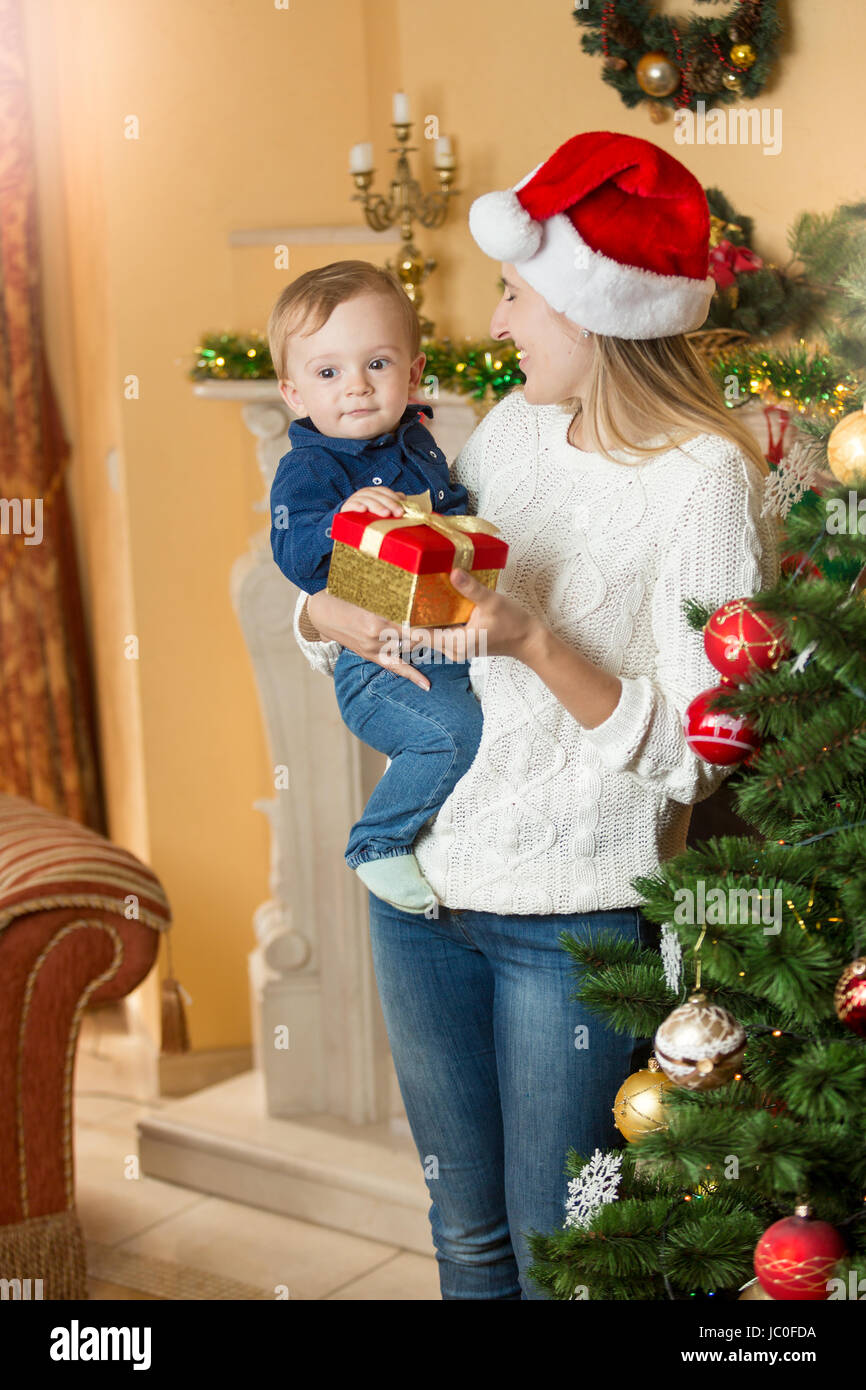 Portrait of happy young mother giving present to her baby son on Christmas at living room next to Christmas tree Stock Photo