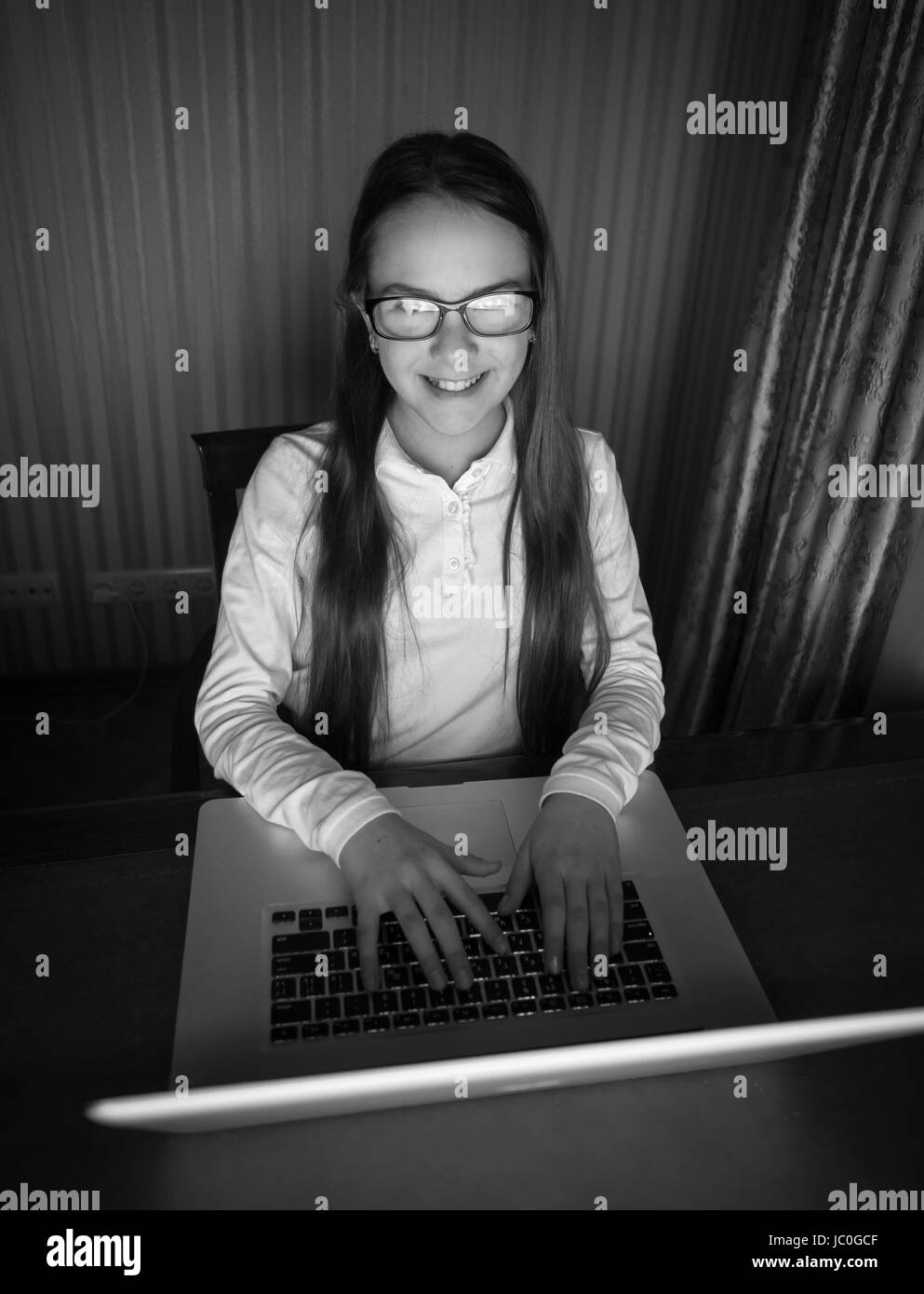 Black and white portrait of teenage girl in eyeglasses sitting at computer in dark room Stock Photo
