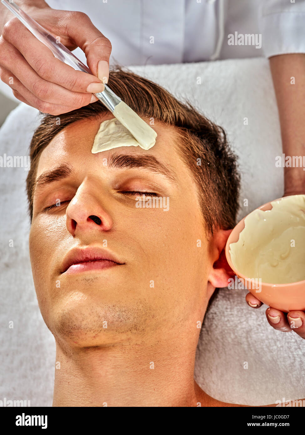 Mud facial mask of man in spa salon. Massage with clay full face. Man lying  on spa bed. Beautician with bowl therapeutic procedure isolated background  Stock Photo - Alamy
