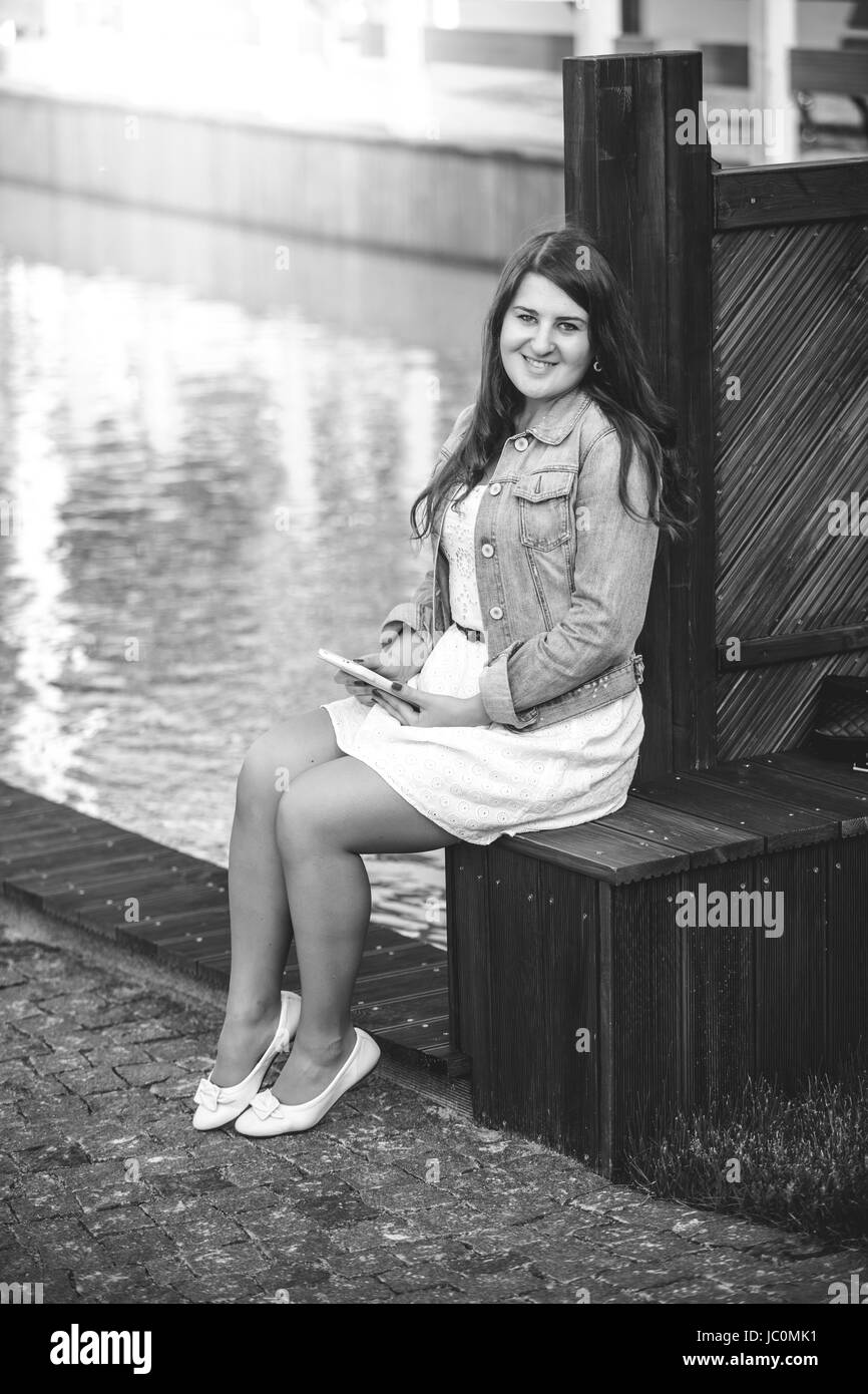Black and white photo of young woman sitting on bench near river with tablet Stock Photo