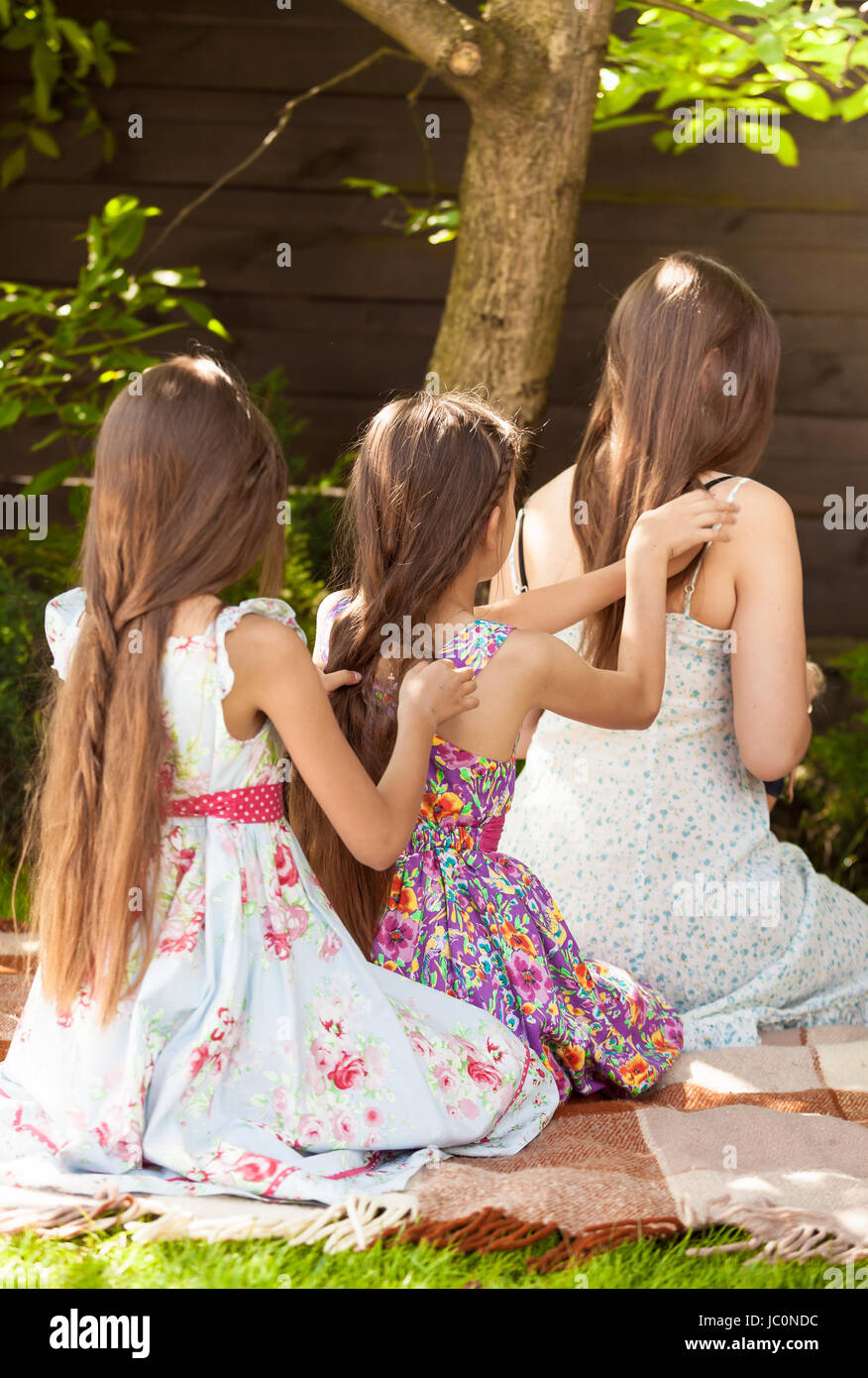 Three girls sitting under tree at park and braiding each other Stock Photo