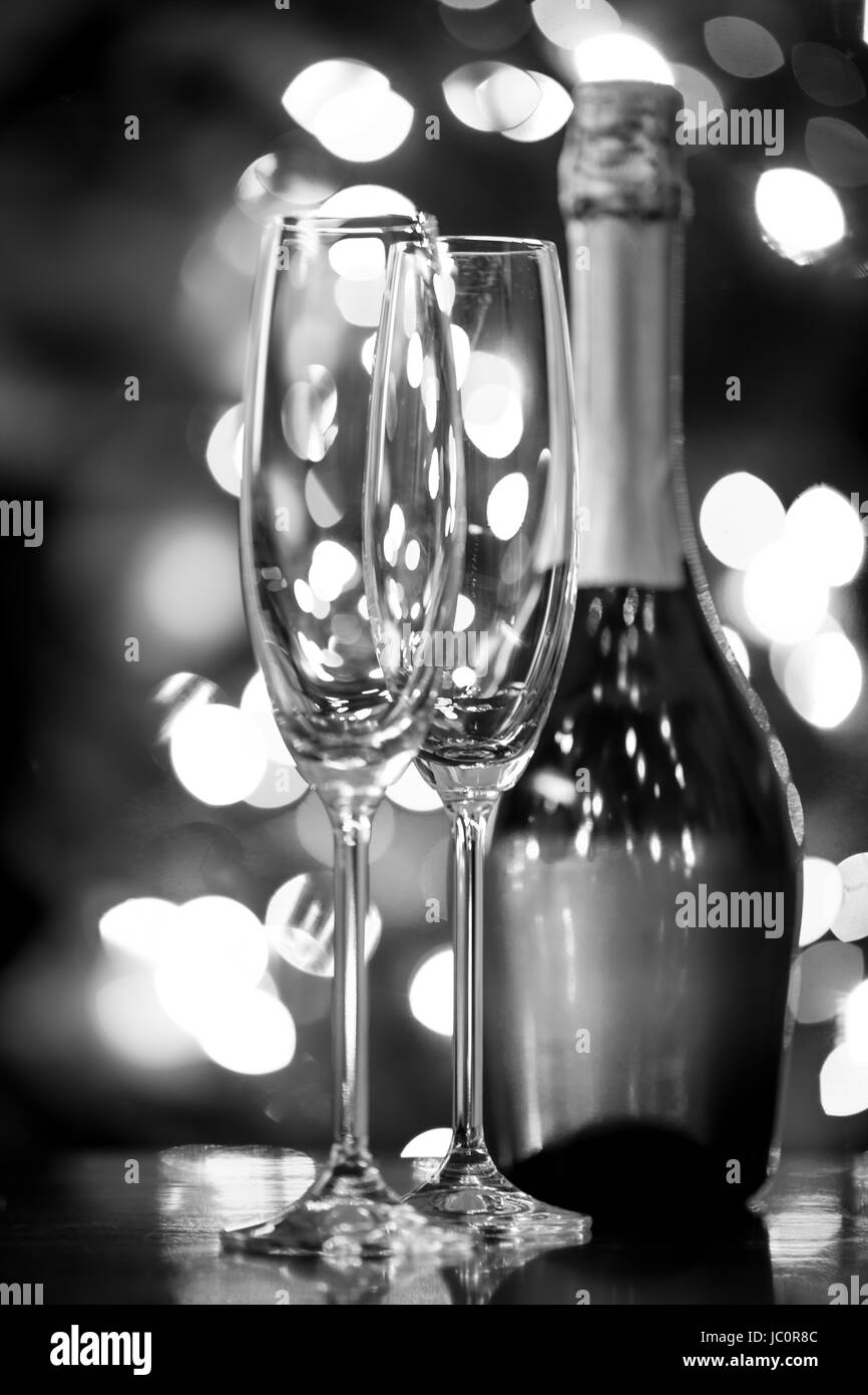 Black and white closeup photo of two glasses and bottle of Champagne against Christmas lights Stock Photo