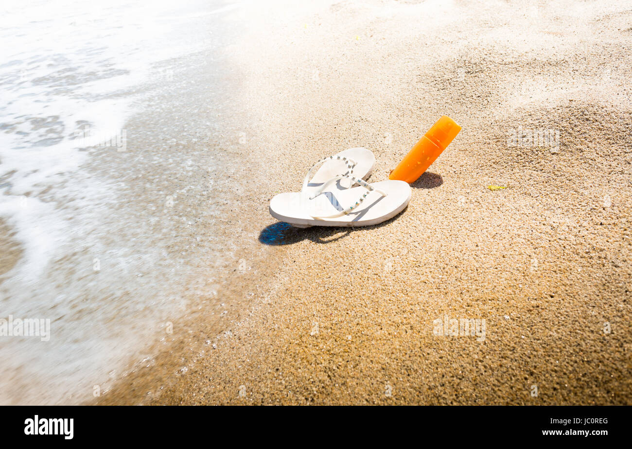 Conceptual shot of flip flops and sunblock lotion on sandy sea beach Stock Photo