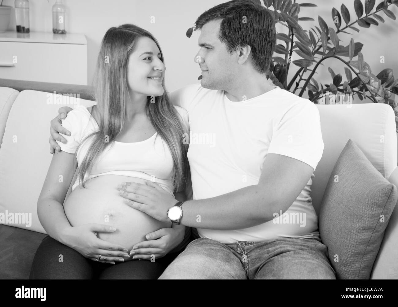 Black and white portrait of happy pregnant couple sitting on sofa and looking at each other Stock Photo