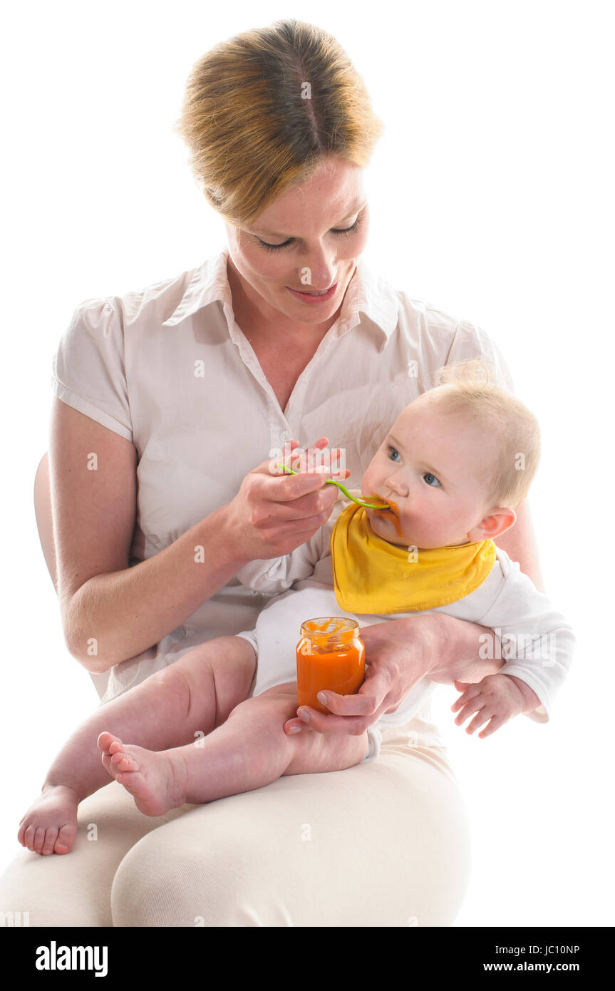 Erwachsene Frau sitzt auf einem weissen Stuhl und haelt das 6 Monate alte Baby im Arm und fuettert mit einem gelben Plastikloeffel einen orangen Karottenbrei, isoliert vor weissem Hintergrund. Stock Photo