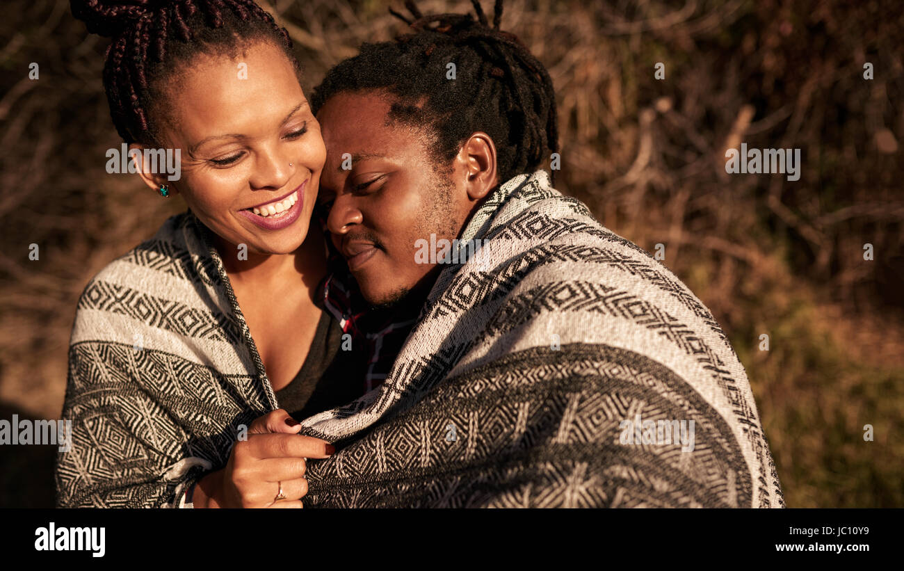 Young mixed race couple embracing under blanket outdoors during sunset Stock Photo