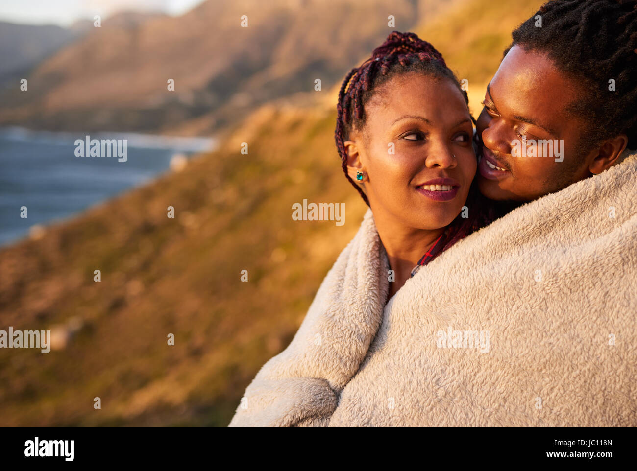 Cute affectionate interracial couple keeping warm with a blanket outdoors Stock Photo