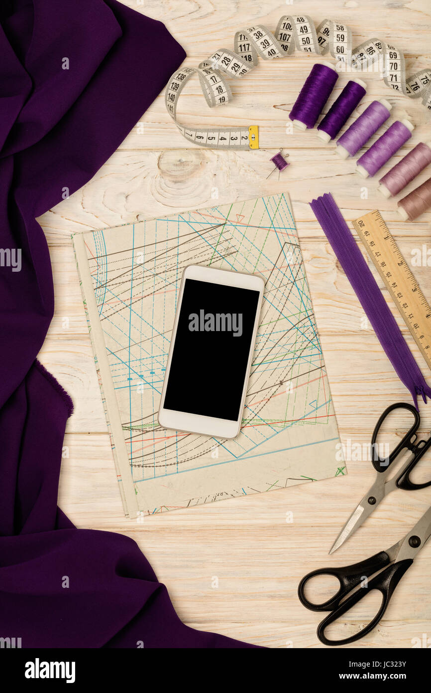 Sewing accessories, fabric, patterns and a mobile phone of white color on a light wooden background. Selective focus. Stock Photo