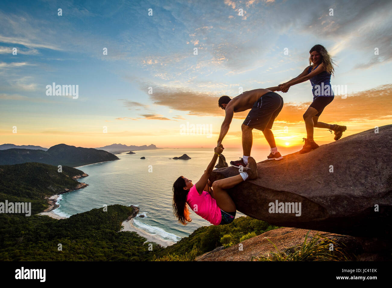 Two people helping woman climb up rock at sunset, Pedra do Telegrafo, Rio de Janeiro, Brazil Stock Photo