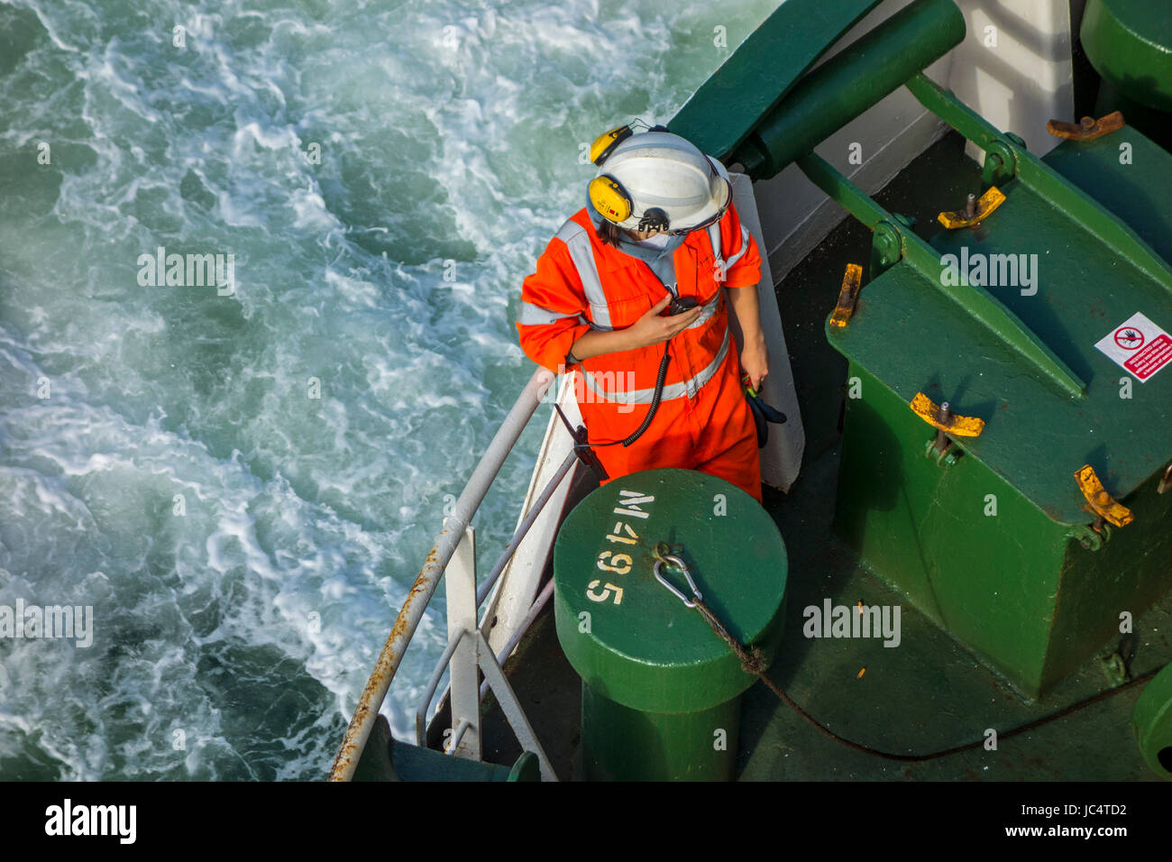 Female crew member with radio / walkie-talkie in orange overall and wearing safety helmet working on deck of car carrier / cargo ship at sea Stock Photo