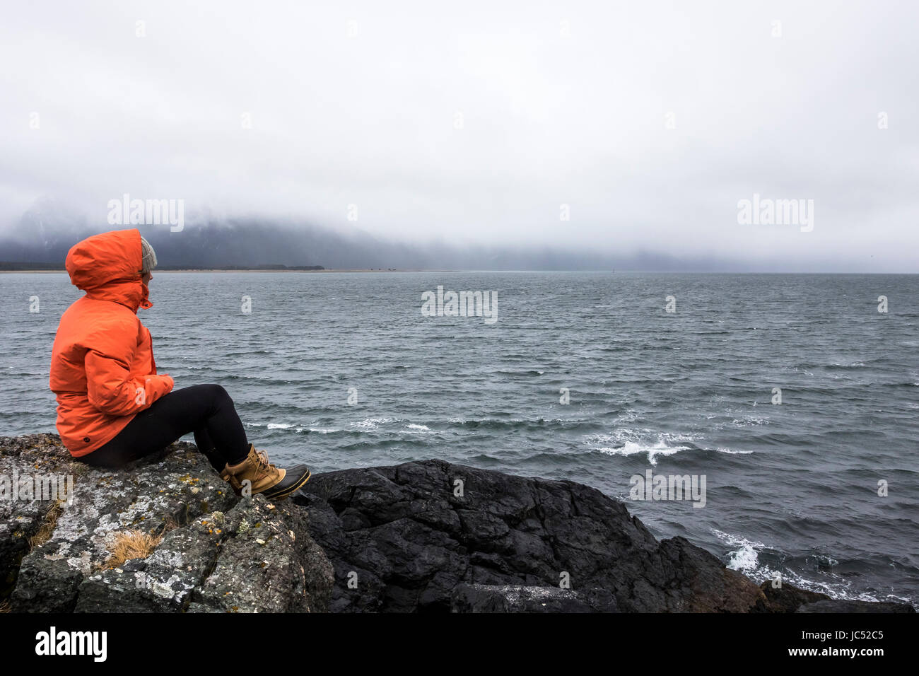 Professional Snowboarder Robin Van Gyn sits on a rock and looks over the ocean on a stormy down day while on snowboard trip to Haines, Alaska. Stock Photo