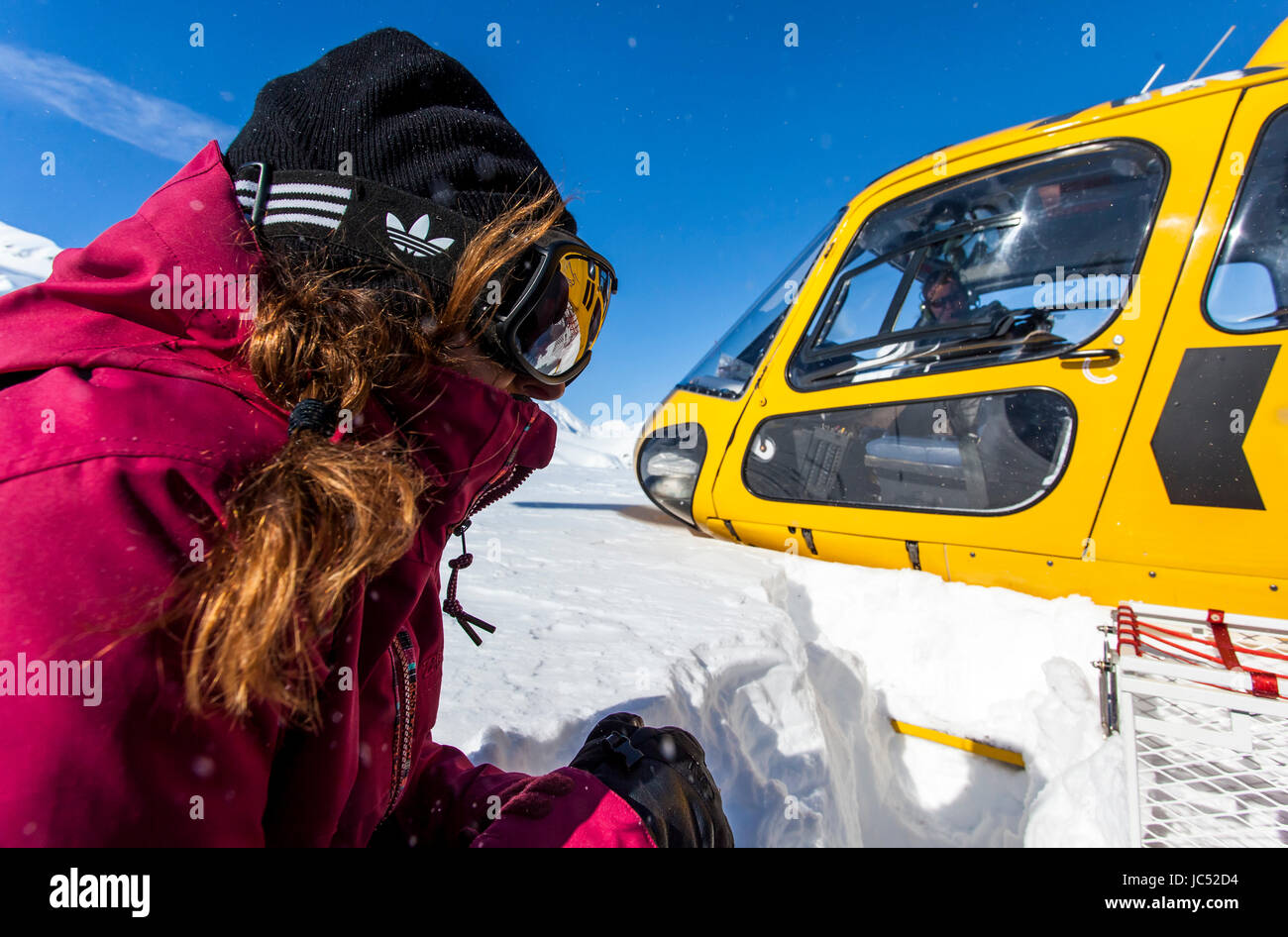 Professional snowboarder Helen Schettini crouches down as a helicopter takes off next to her on a sunny blue bird day in Haines, Alaska. Stock Photo