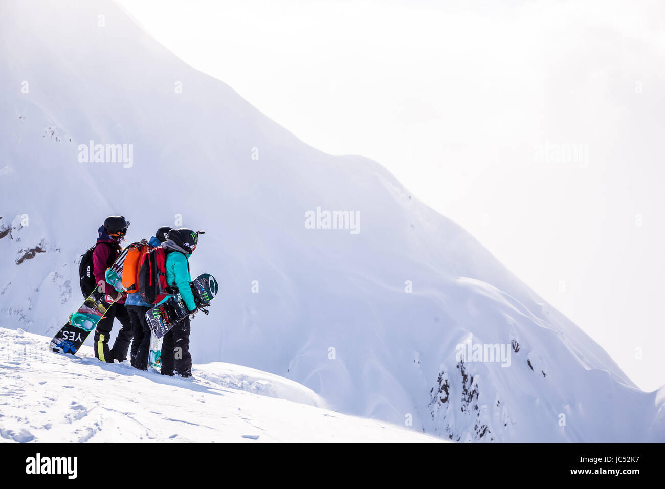 Professional snowboarders Robin Van Gyn, Helen Schettini, and Jamie Anderson, stand on a ridge and look down at a line they are about to ride on a sunny day in Haines, Alaska. Stock Photo