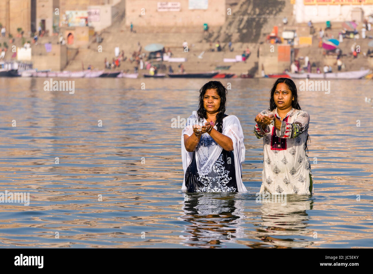 Two women, pilgrims, are taking bath and pray on the sand banks at the holy river Ganges, panorama of Dashashwamedh Ghat, Main Ghat, in the distance Stock Photo