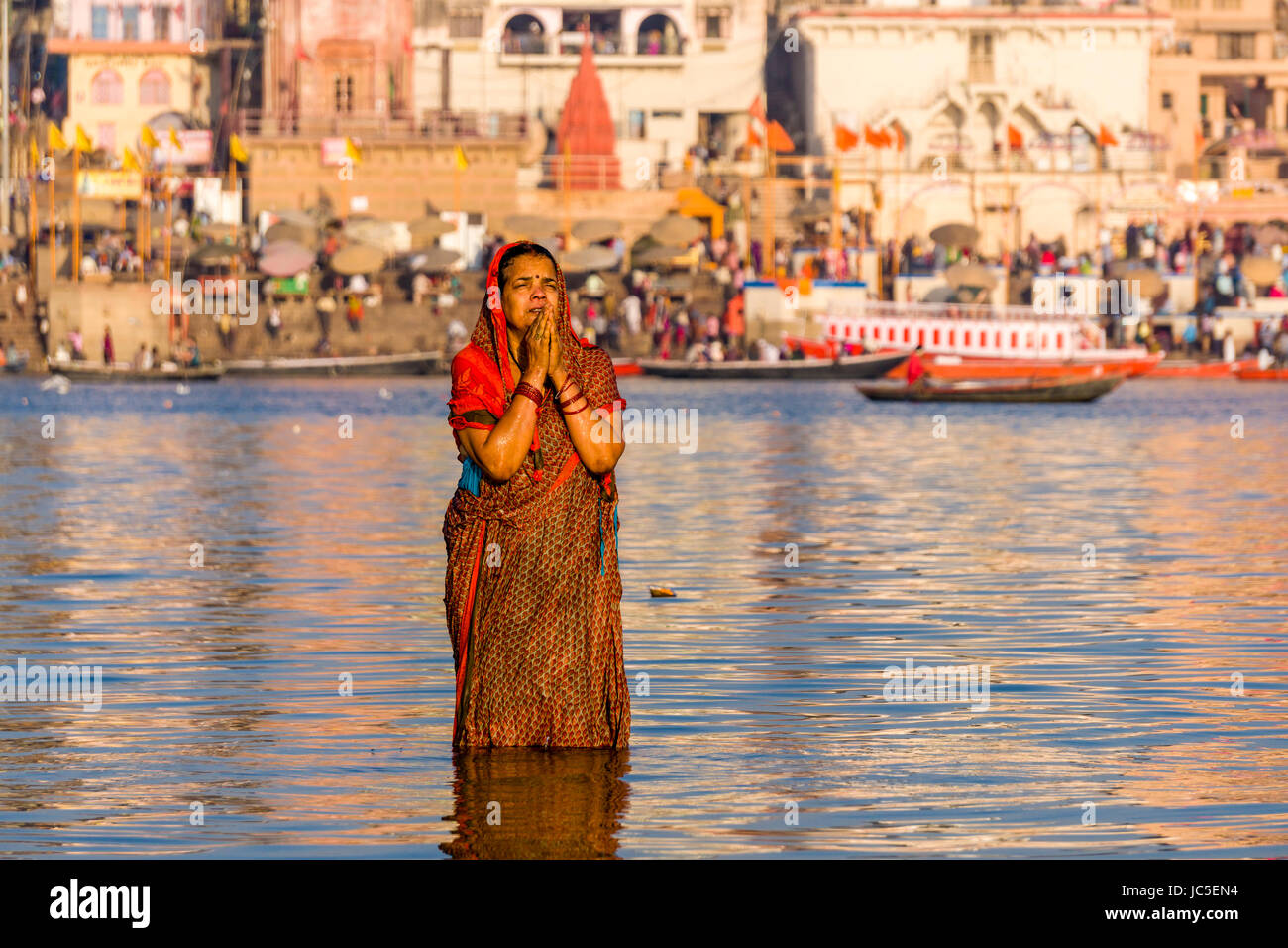 A woman, pilgrim, is taking bath and praying on the sand banks at the holy river Ganges, panorama of Dashashwamedh Ghat, Main Ghat, in the distance Stock Photo