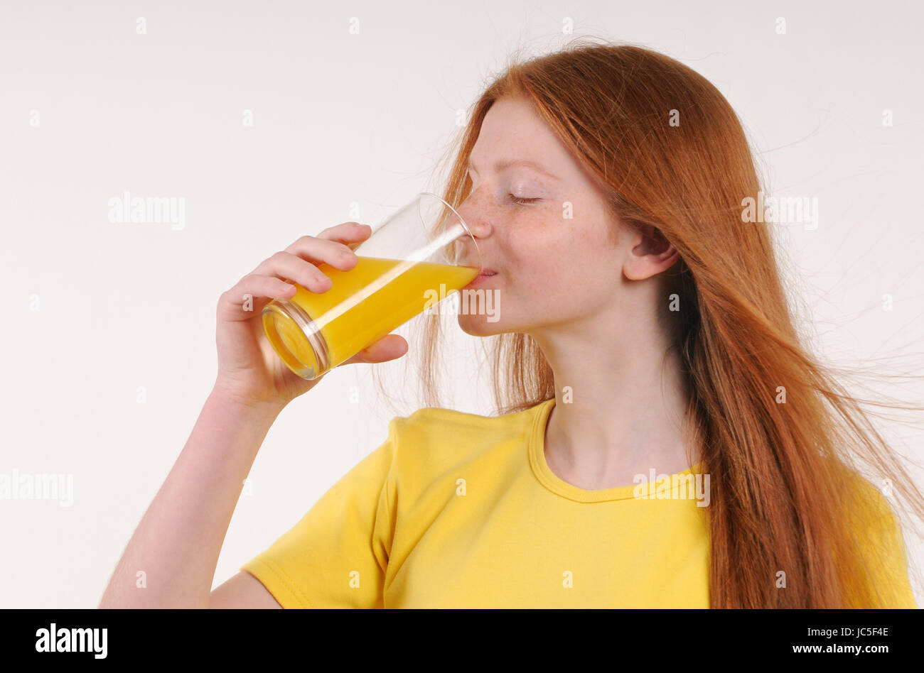 Red hair teenage girl drink orange juice with glass. Gray isolated portrait drinking girl. Stock Photo