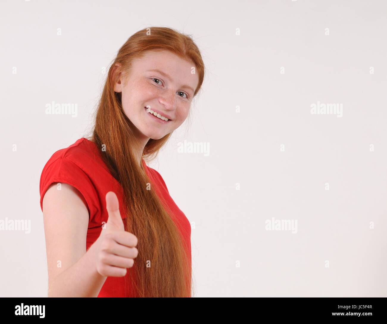 Red hair tennager girl in a red shirt showing a thumbs-up on grey background. Happy smiling lifestyle people concept. Human emotions. Stock Photo