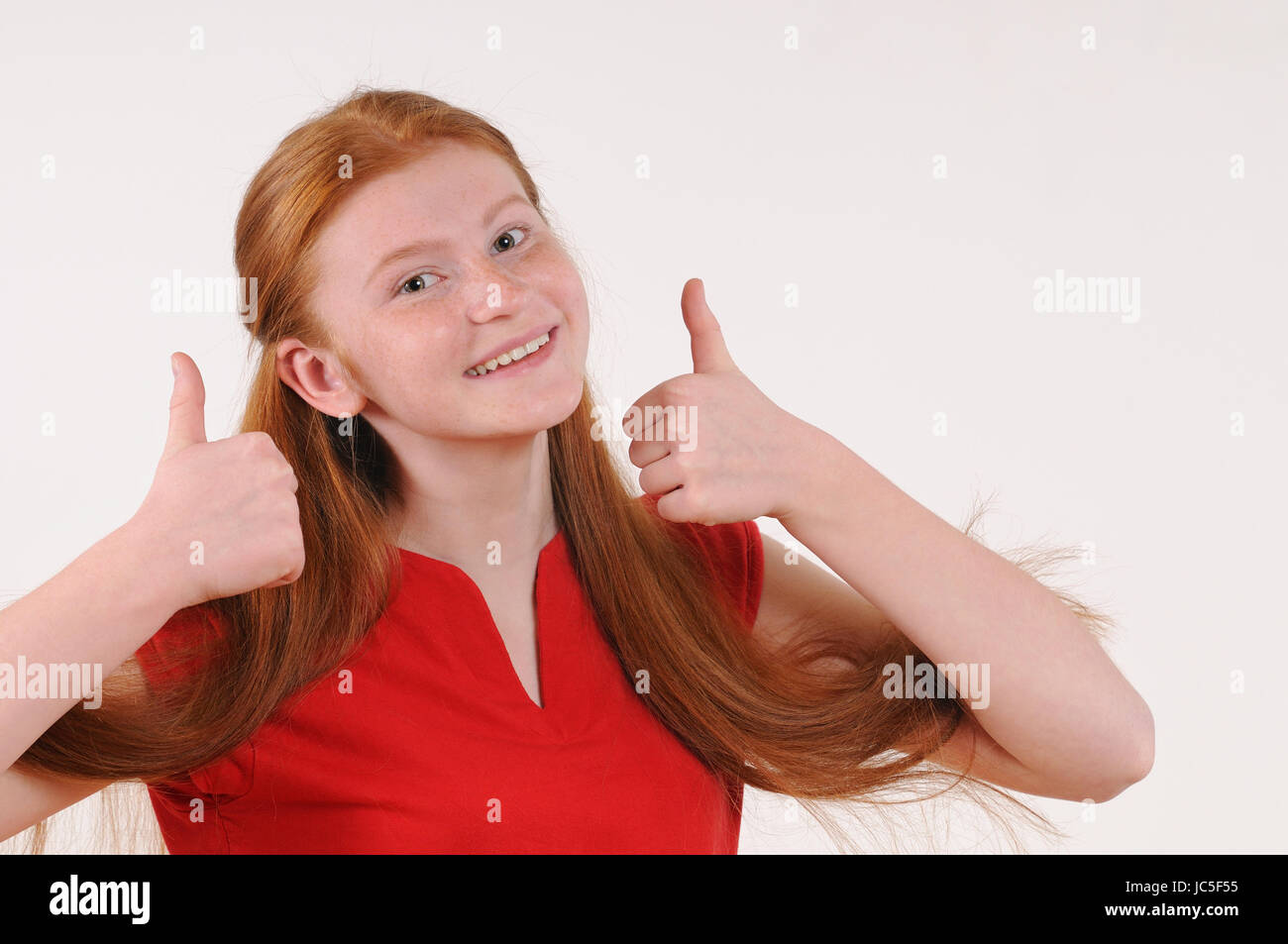 Red hair tennager girl in a red shirt showing a thumbs-up on grey background. Happy smiling lifestyle people concept. Human emotions. Stock Photo