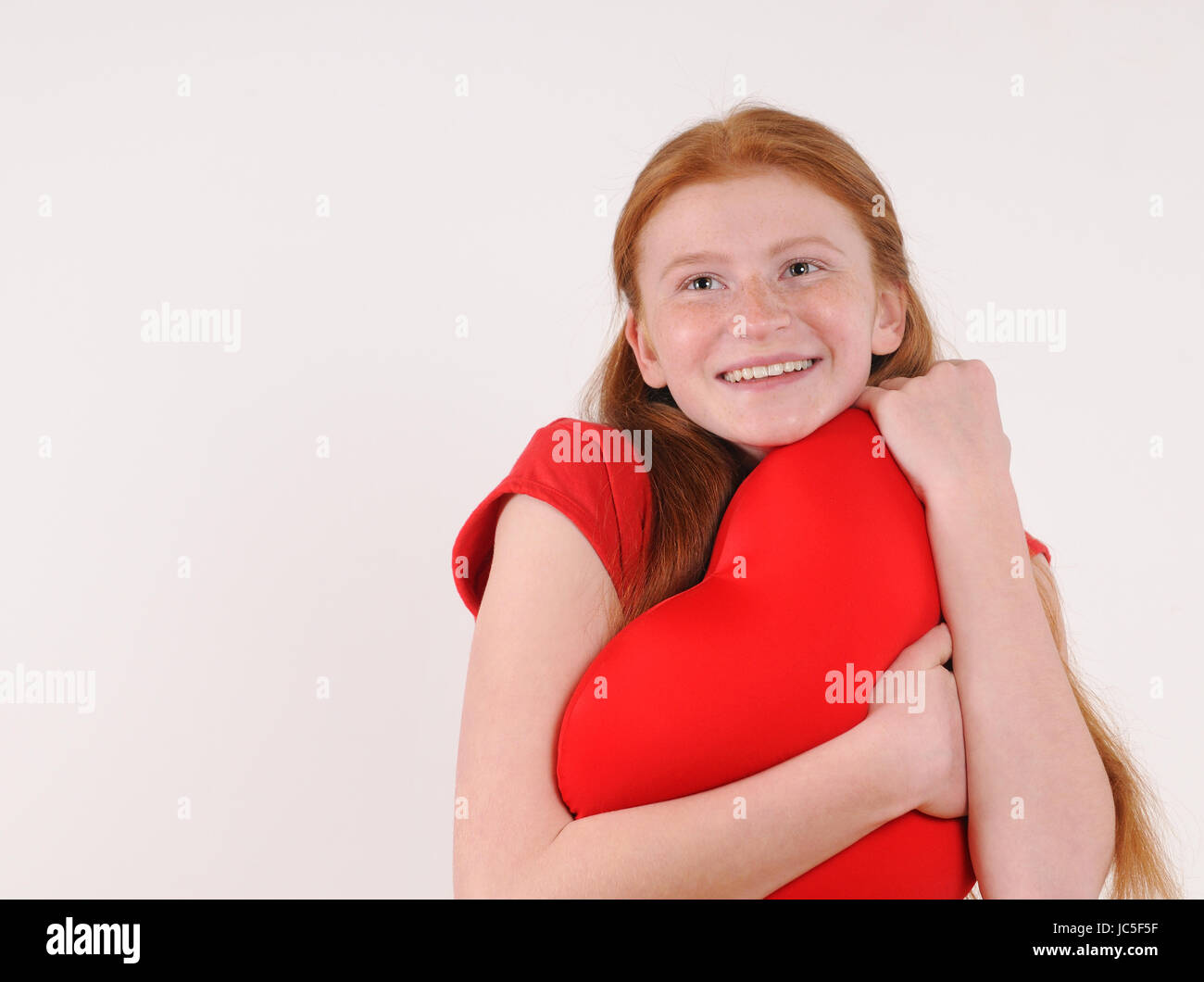 Young red hair girl tenderly hug a heart shape on grey background. Happy smiling lifestyle people concept. Human emotions. Stock Photo