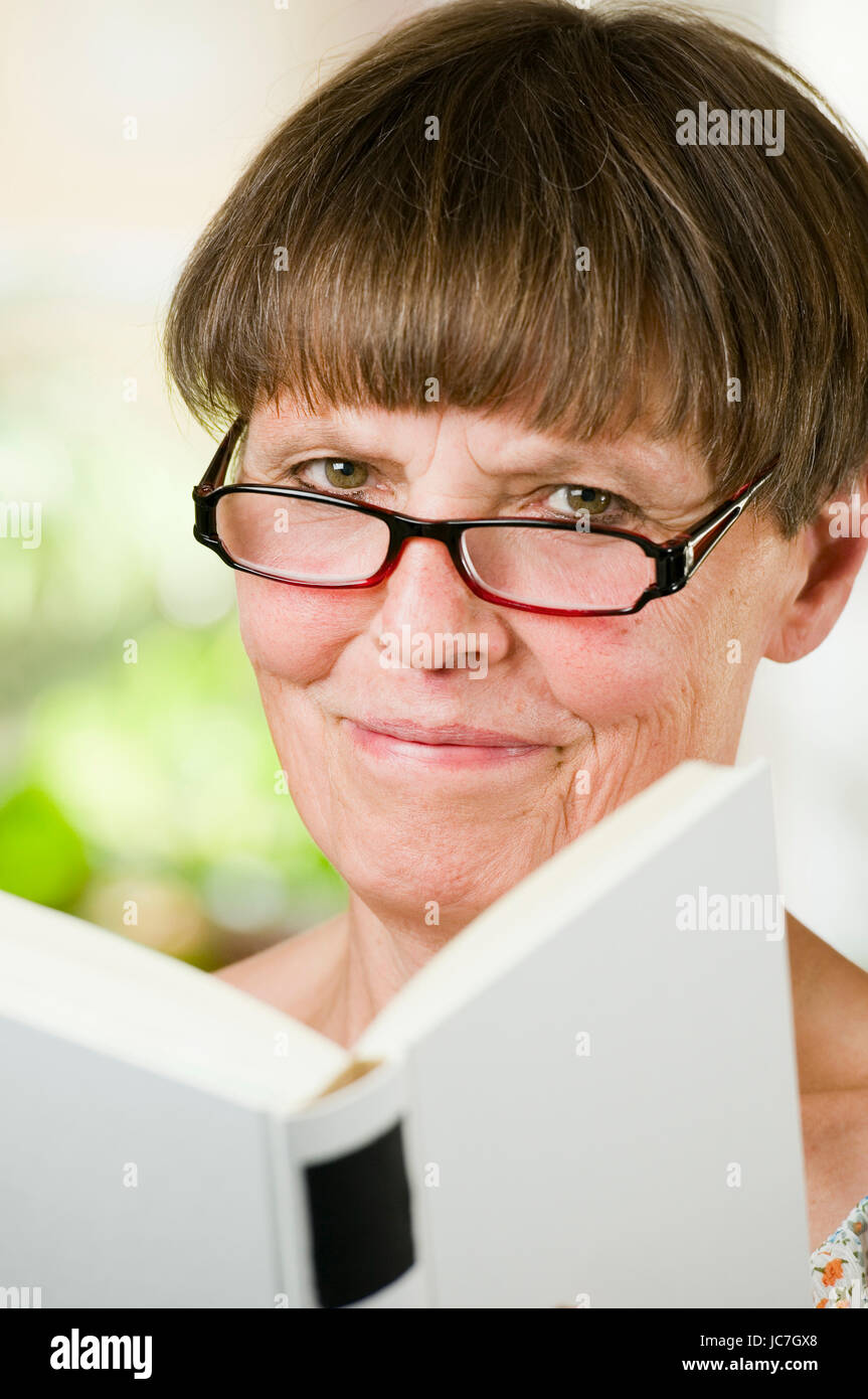 Innenaufnahme Kopf-und-Schulter-Porträt einer älteren Frau mit Lesebrille und offenem buch freundlich in die Kamera lächelnd Stock Photo