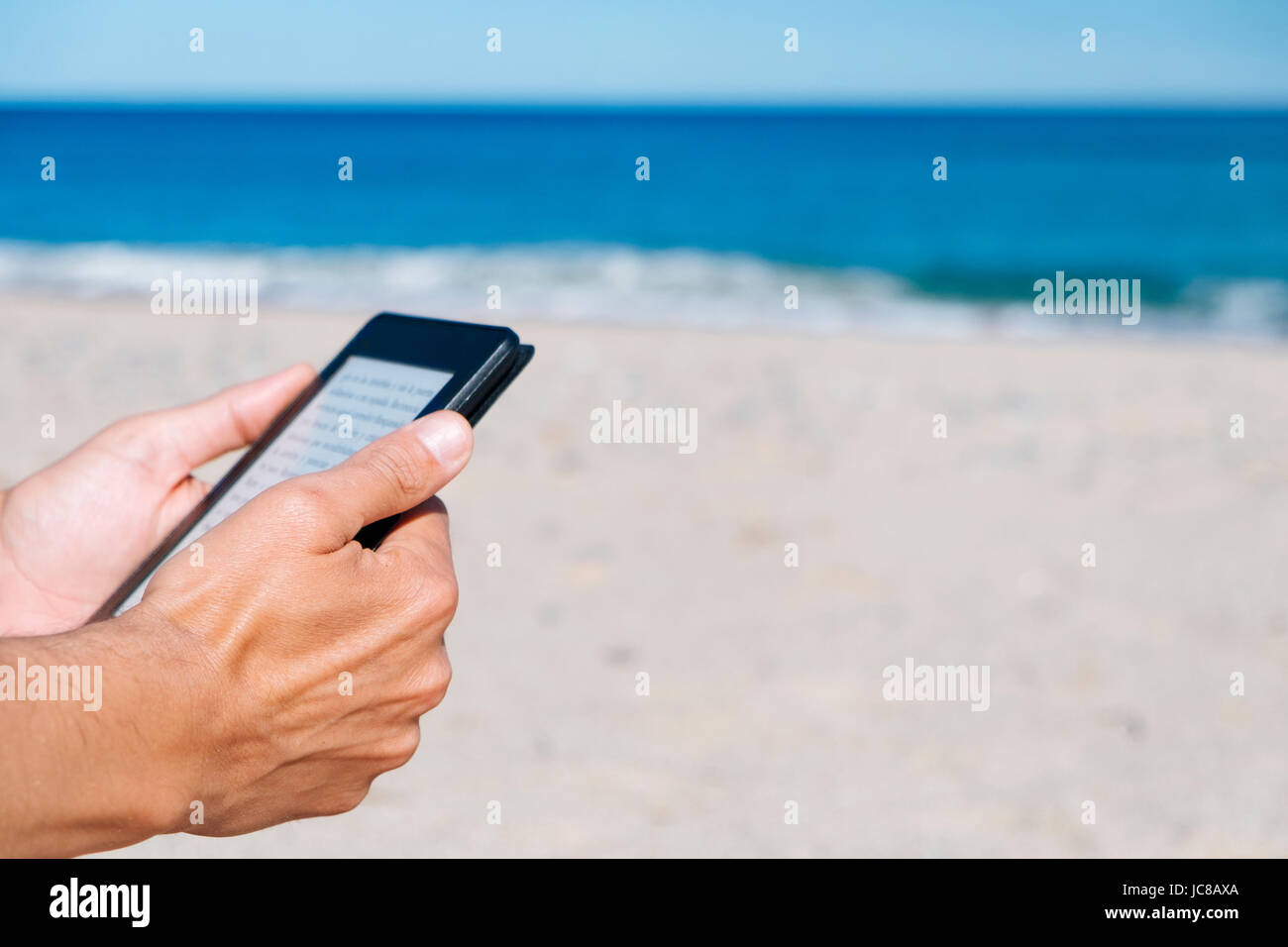 closeup of a young caucasian man reading in a tablet or e-reader on a white sand beach next to the sea Stock Photo