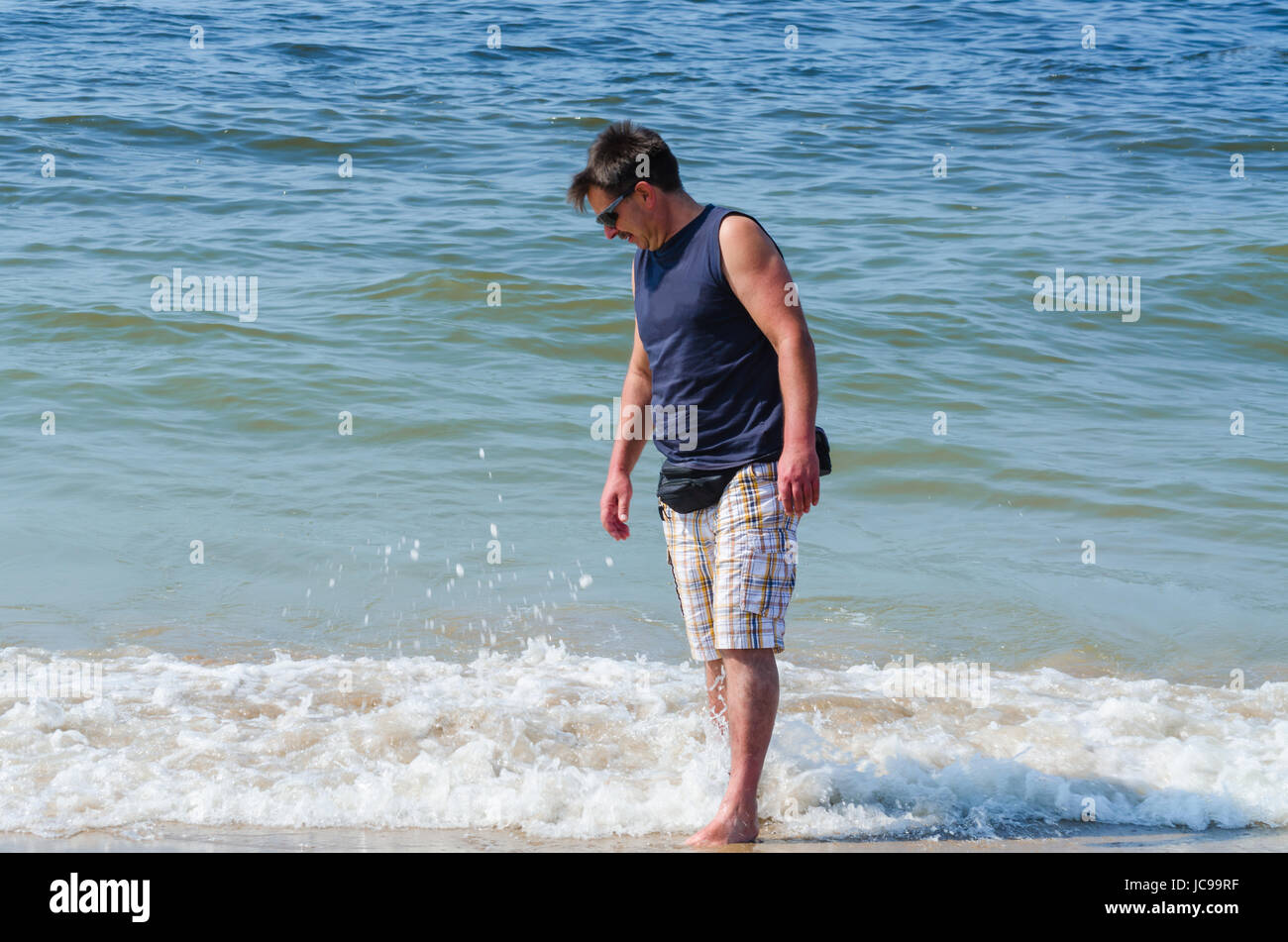 Mann in Shorts, T-Shirt und Brille am Strand  mit den Füßen im Wasser. Stock Photo