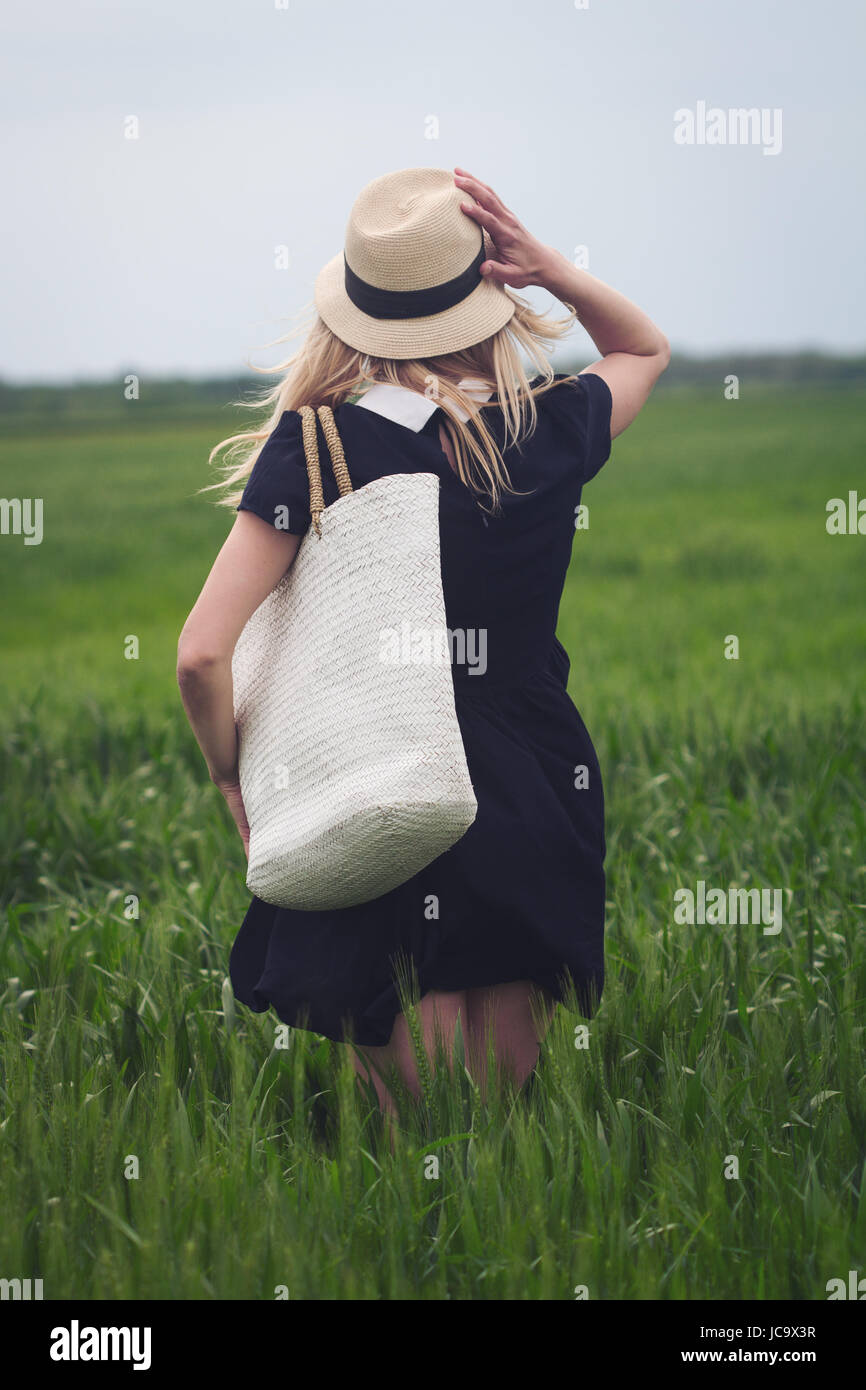 rear view of beautiful Caucasian woman with wicker hat and white bag wearing a black dress and enjoying nature in a green field of wheat Stock Photo