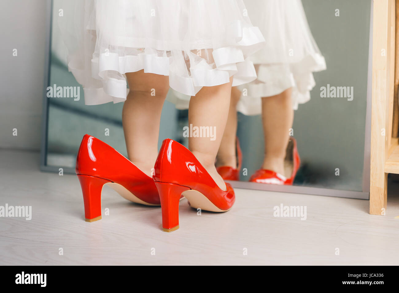 Cute funny little baby girl walks at home trying mom's red high heel shoes Stock Photo