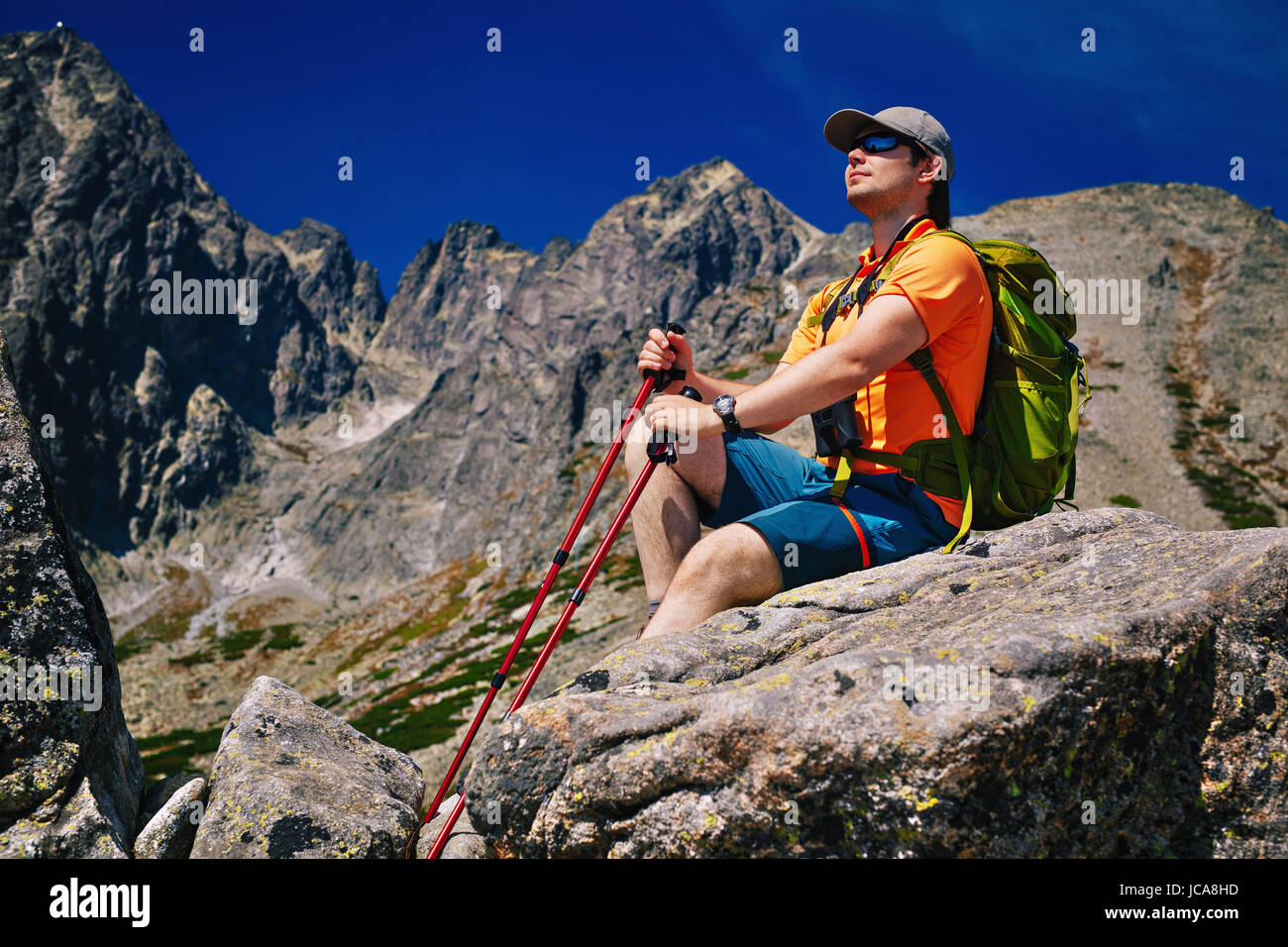 Young man tourist with green backpack and poles sitting at high mountains and looking aside Stock Photo
